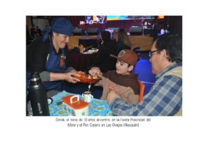 "El niño Derek aparece sentado en el centro de una mesa con un equipo de mate. A la izquierda, una mujer mayor con un pañuelo azul en la cabeza le ofrece bizcochos en una bandeja pequeña. A la derecha, un hombre está sentado junto a él, durante la Fiesta Provincial del Mate y el Pan Casero